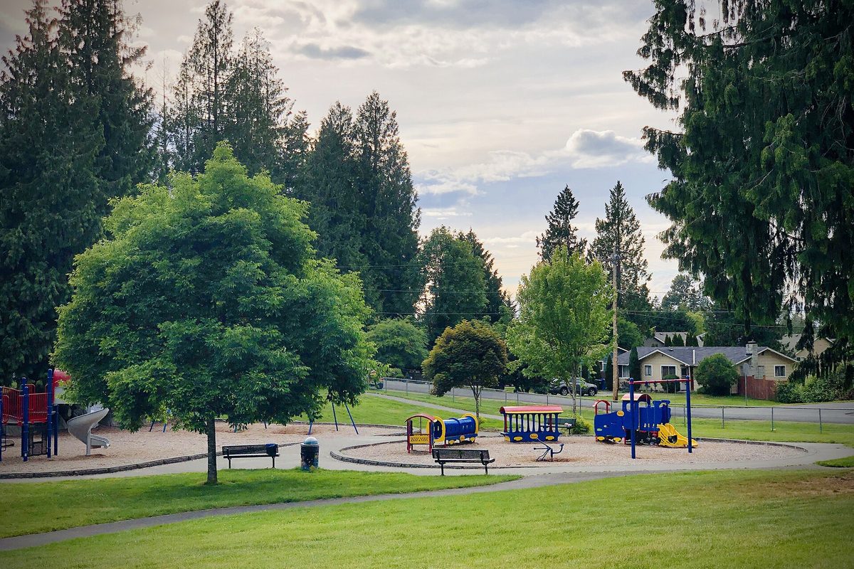 Playground with a blue toy train, picnic benches and lawn in Cedarview Park, Bonney Lake, with tall evergreens and houses behind