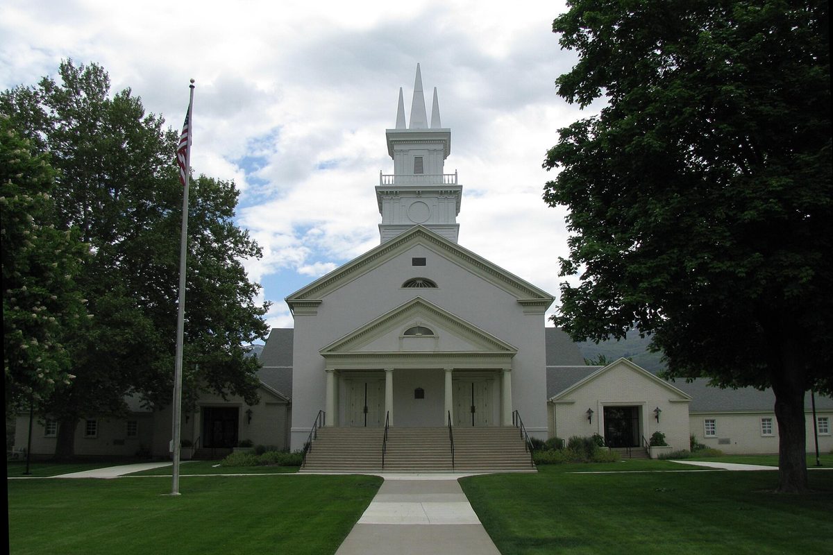 The white Bountiful Tabernacle with columns, wide front steps and a tall steeple, between big shade trees and a flagpole