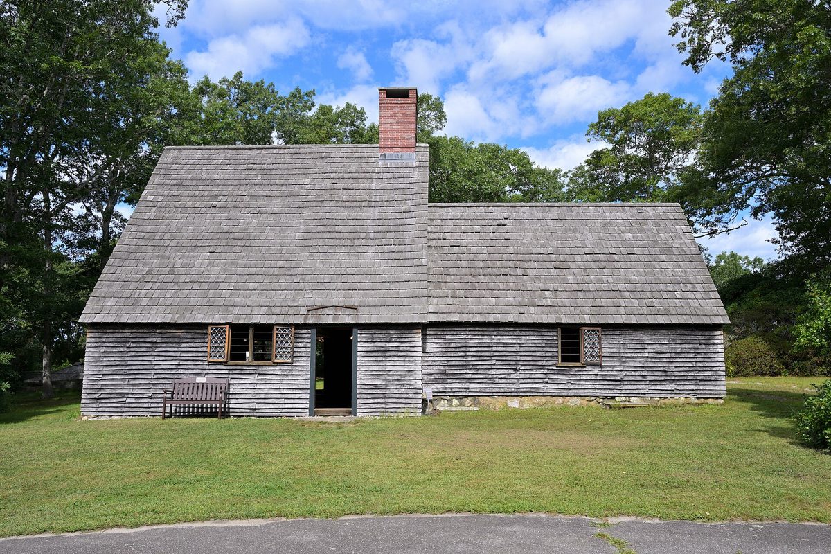 The weathered gray-shingled replica of the Aptucxet Trading Post in Bourne, with a tall brick chimney, an open door and a bench on the lawn