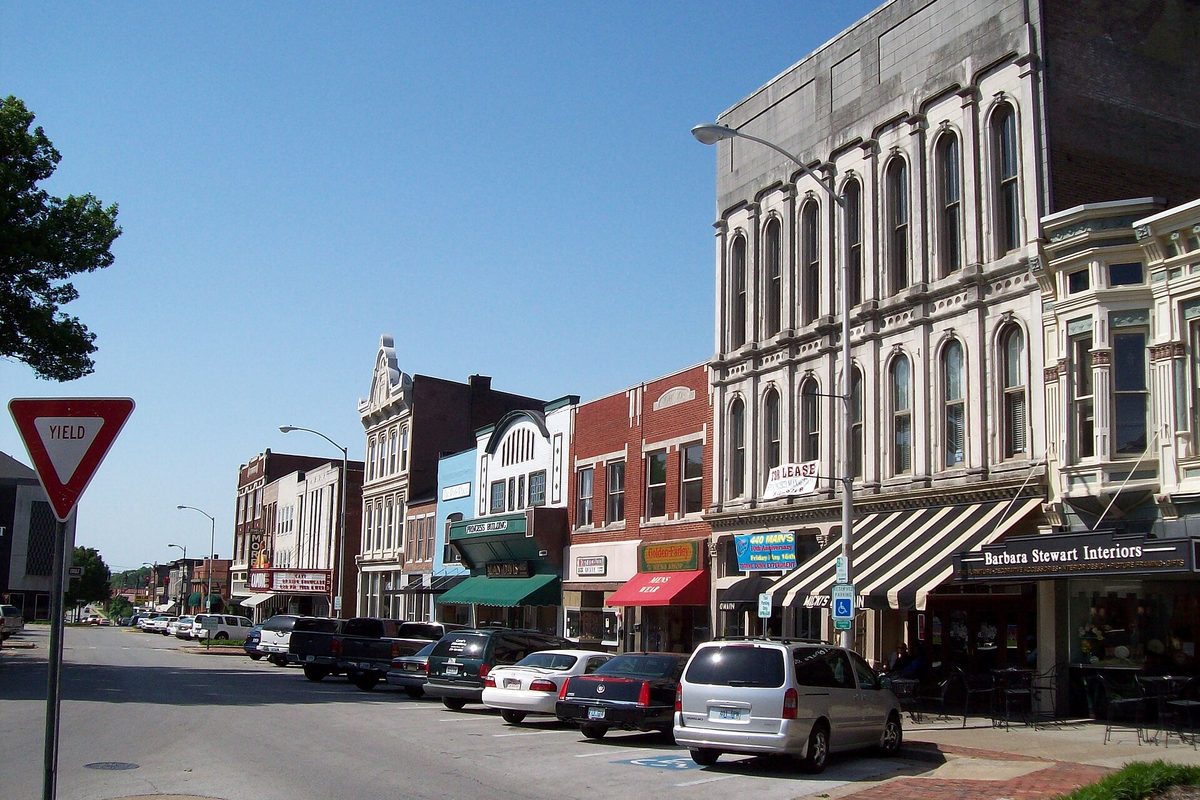 Brick and stone storefronts with awnings along Fountain Square in downtown Bowling Green, cars parked at the curb and a yield sign in the foreground