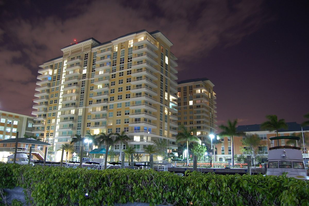 The Marina Village condo towers in Boynton Beach lit up at night, with palm trees, a docked boat and a hedge in the foreground