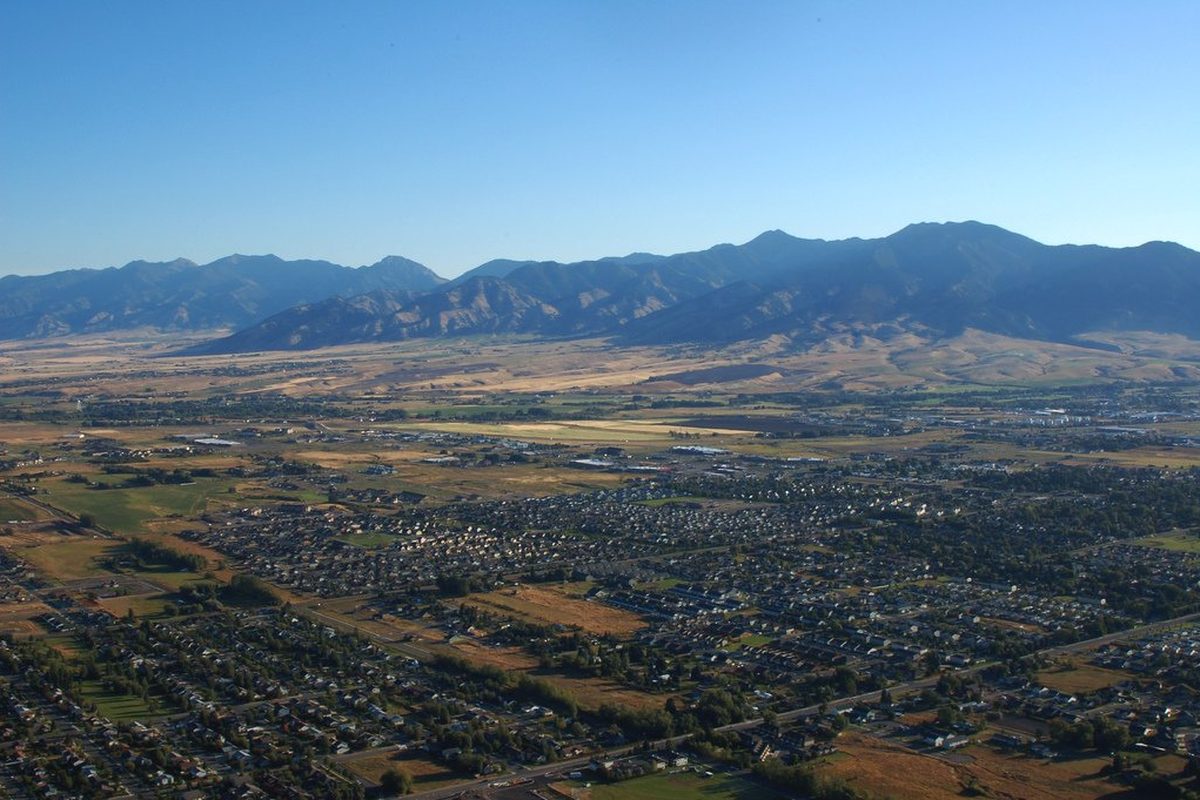 Aerial view of Bozeman's neighborhoods and open fields stretching across the valley floor toward a dark mountain range under a clear blue sky