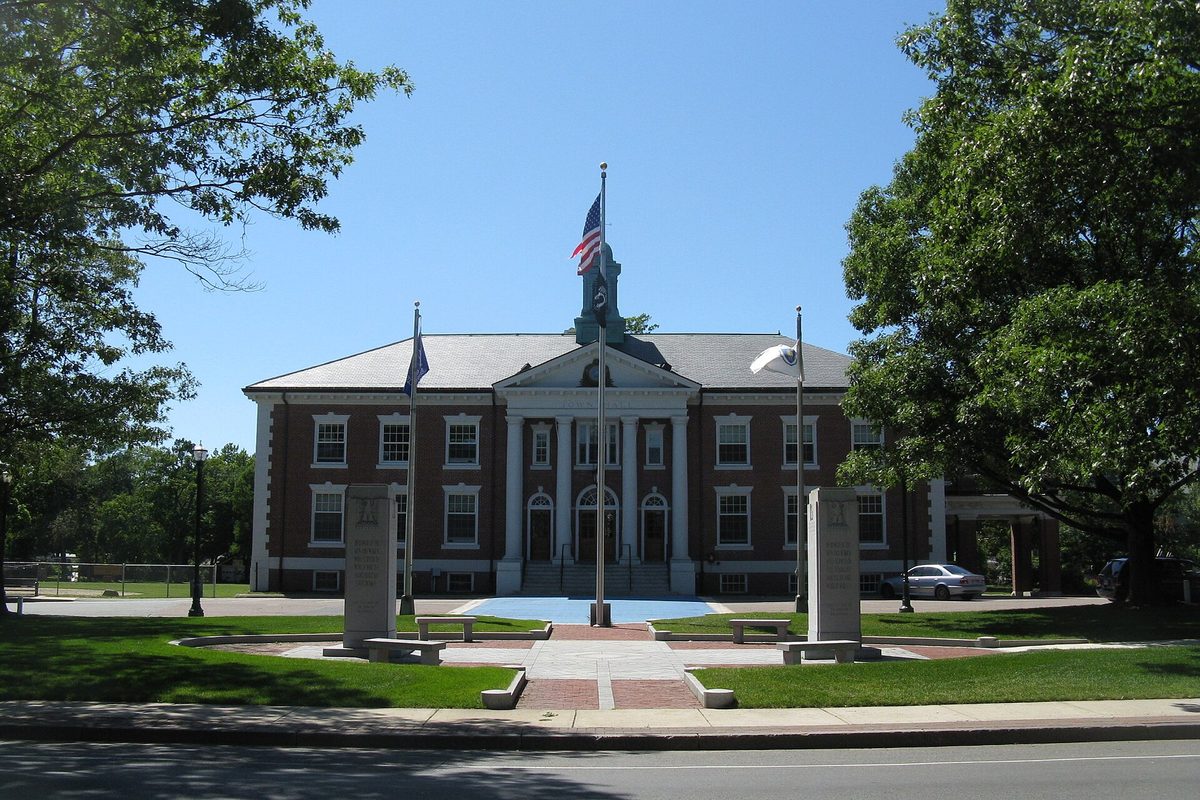 Braintree Town Hall, a red brick building with a white-columned entrance and a small tower, behind a flagpole, stone markers and a green lawn on a sunny day