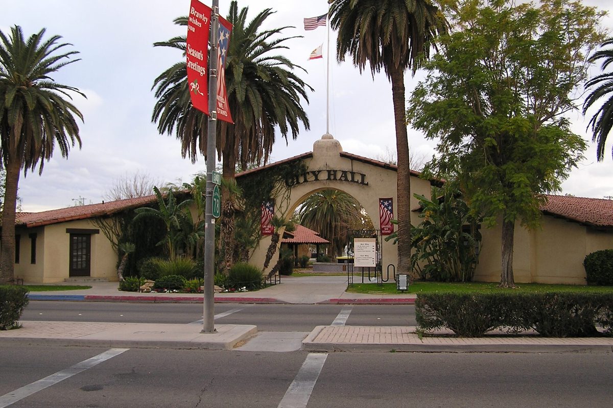 Brawley City Hall, a tan stucco building with a red tile roof and an arched entrance lettered City Hall, framed by palm trees across the street