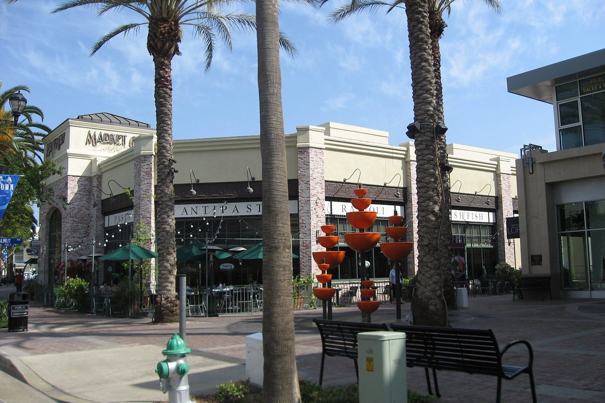 A downtown Brea restaurant storefront with a Market sign and outdoor tables, framed by tall palms, with a bench and orange stacked-bowl fountain sculptures on the plaza