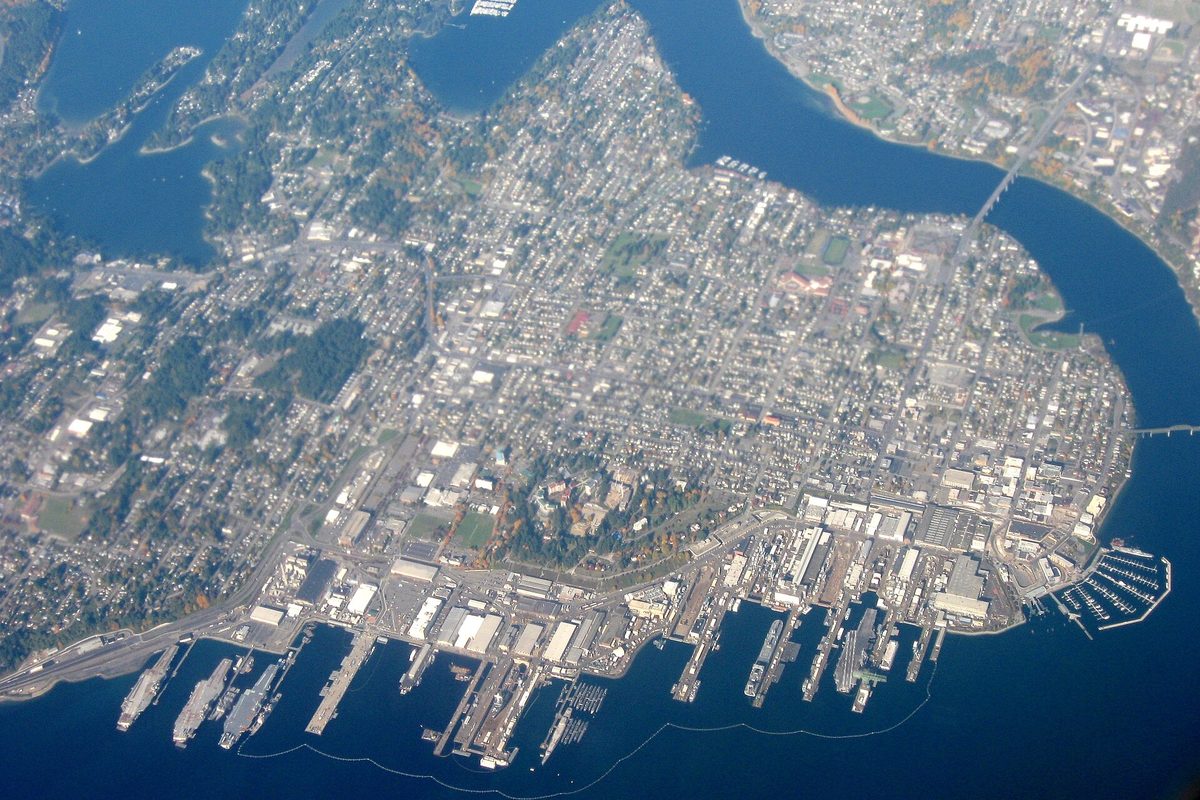 Aerial view of Bremerton on its peninsula, with water on three sides, long piers and docked ships along the south shore and a bridge at the right