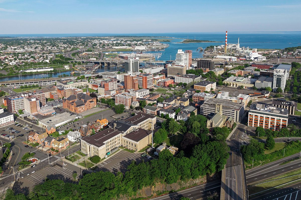 Aerial view of downtown Bridgeport on a summer day, with brick blocks, office towers and highway ramps in front of the harbor and a tall red-and-white smokestack by the water