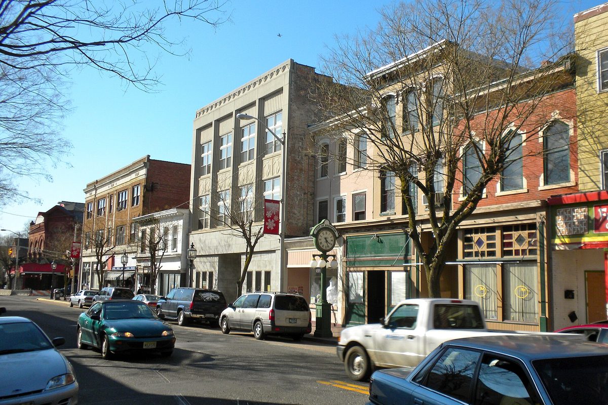 Three- and four-story brick and stone storefronts on Commerce Street in downtown Bridgeton, with a green street clock, bare trees and parked cars under a blue sky