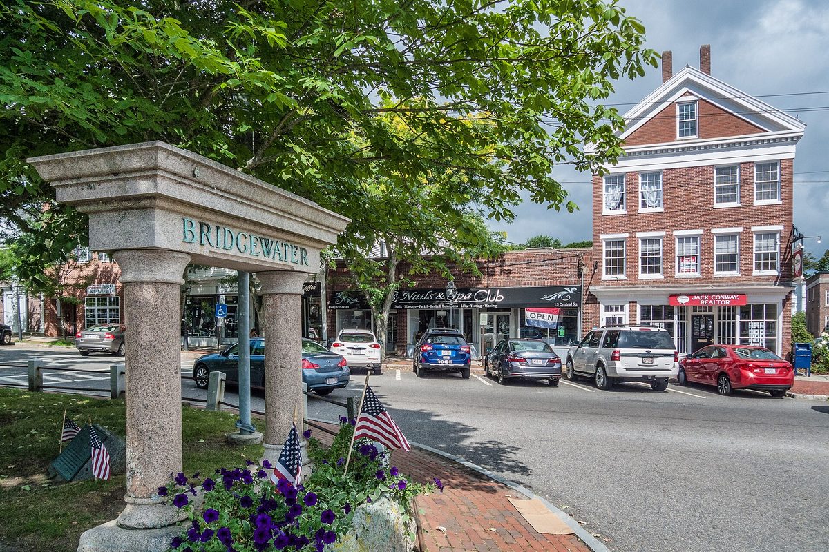 A granite Bridgewater marker with flags and purple flowers in Central Square, with brick buildings, shops and parked cars behind