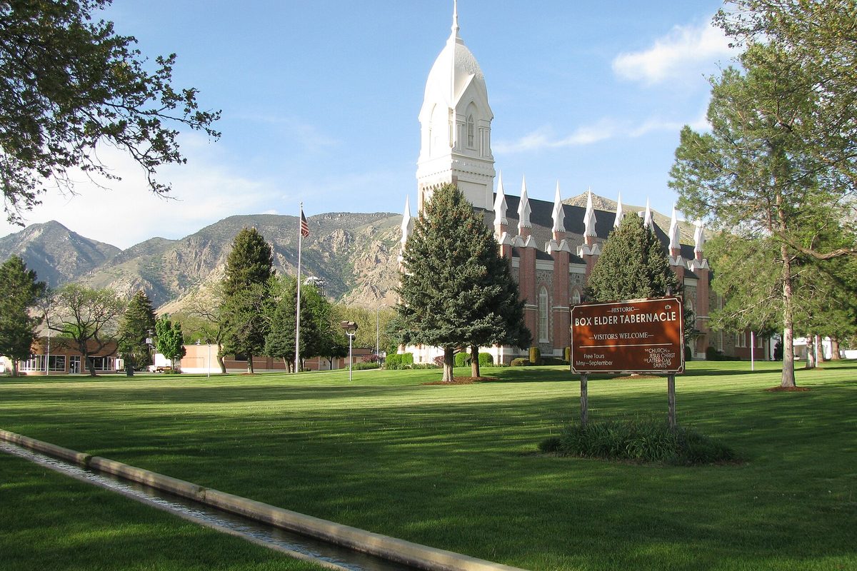 The white-spired Box Elder Tabernacle behind a wide lawn and its brown sign, with rocky mountains rising behind it