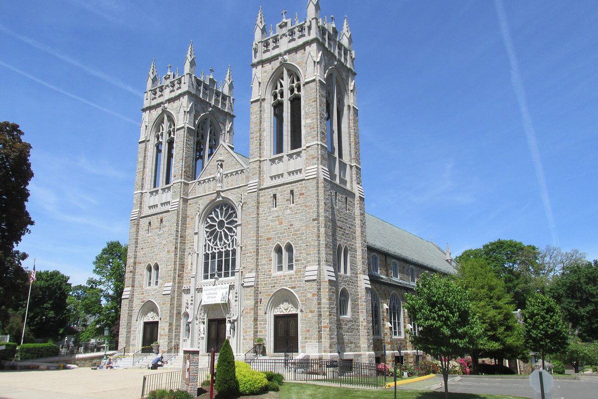 St. Joseph Church in Bristol, a grey stone church with two square Gothic towers, a rose window over the arched doors and trees on both sides under a blue sky