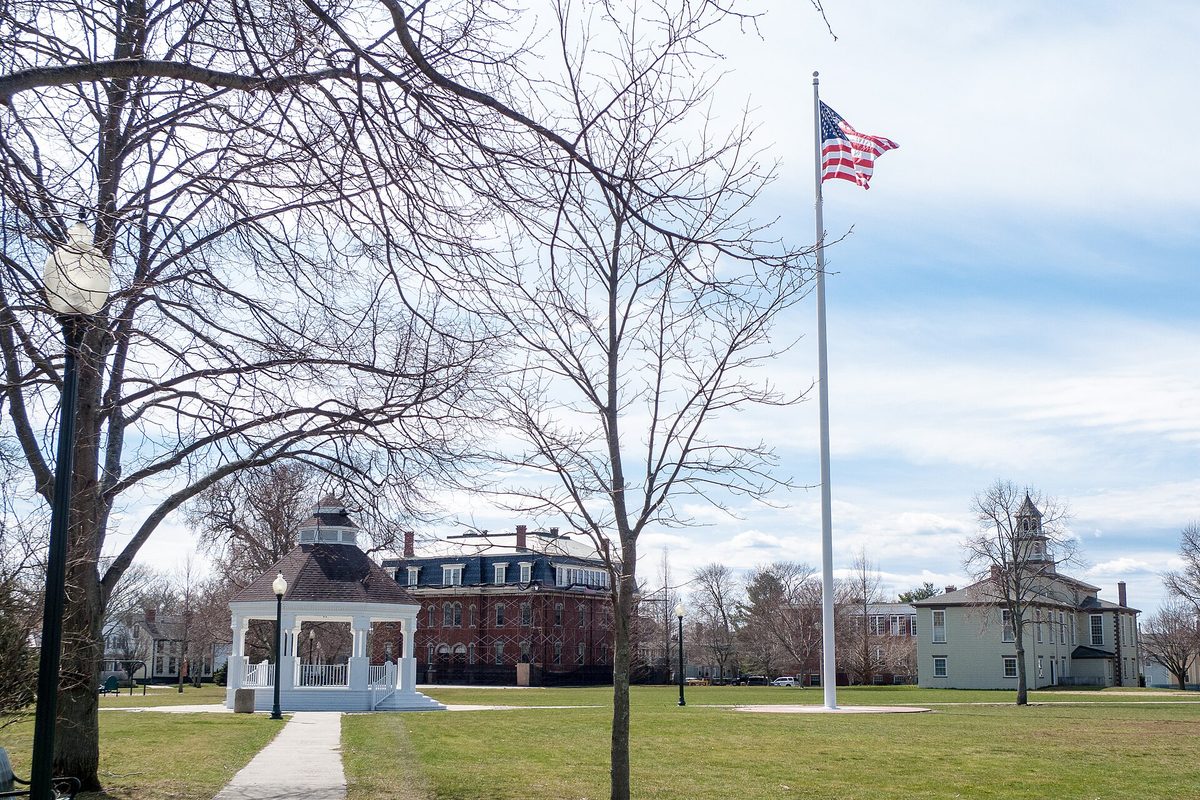 Bristol's town common in early spring, with a white bandstand, a tall flagpole flying the US flag, bare trees and old brick and pale school buildings behind