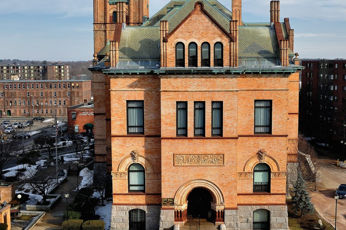 Brockton City Hall, an ornate brick and stone building with a green copper roof and arched entrance, seen from above with snow on the ground nearby