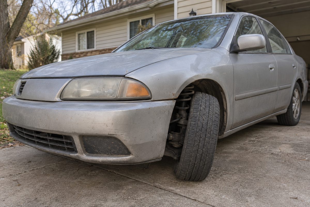 A silver sedan parked in a driveway, sitting low at the front left corner with the tire tucked into the wheel arch and the coil spring showing