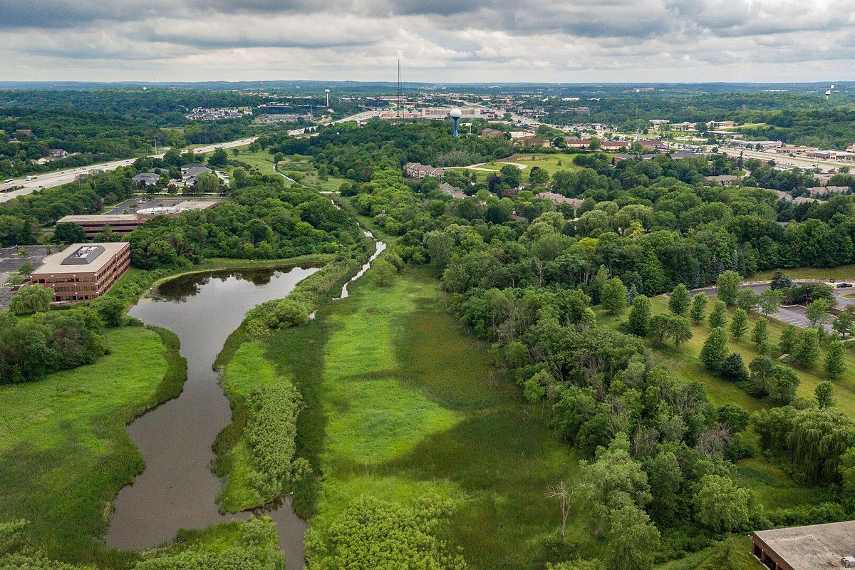 Aerial view over Brookfield of a winding pond and green wetland corridor between office buildings and woods, under grey clouds