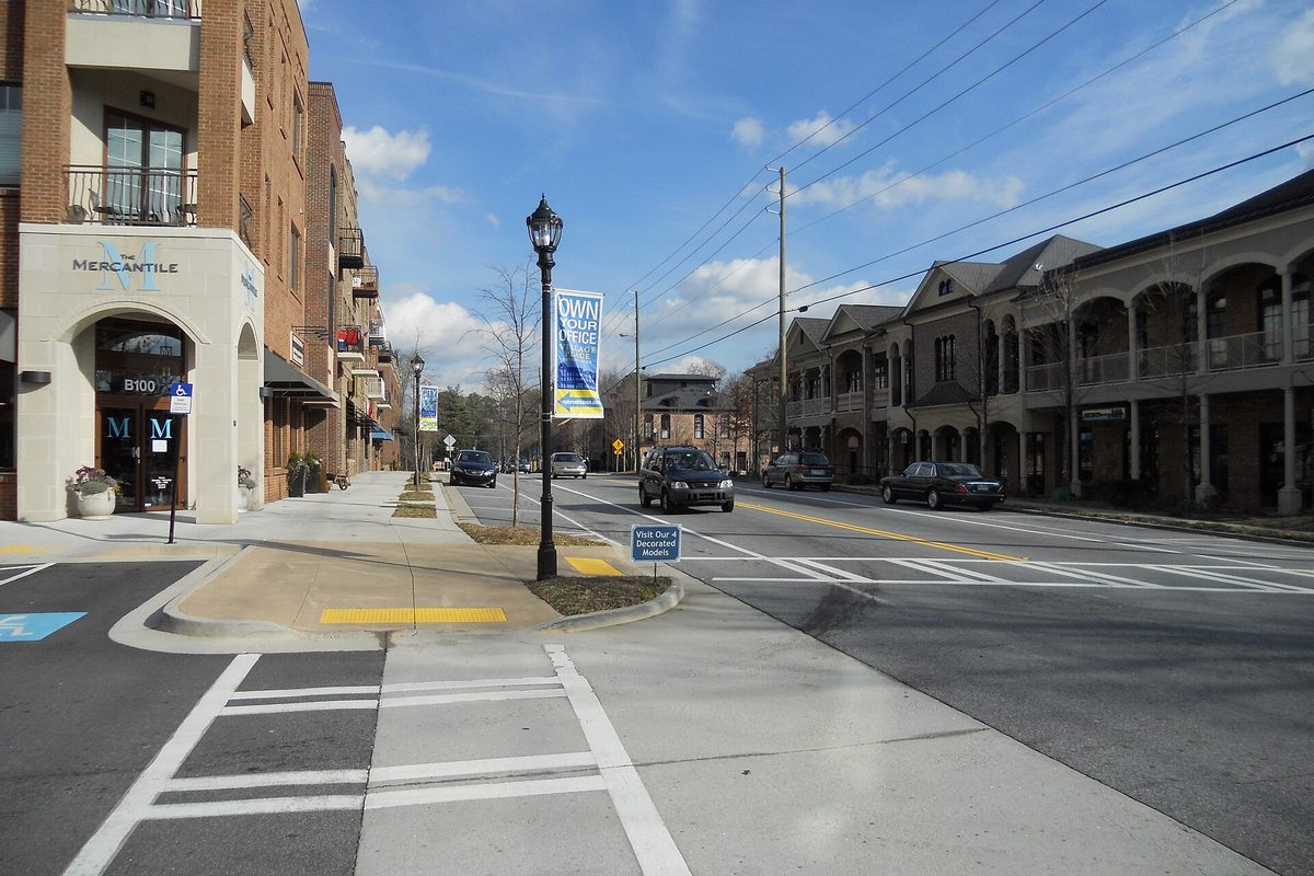 A street corner in Brookhaven Village with a brick shop building, a black lamppost with banners and two-story townhomes across the road under a blue sky