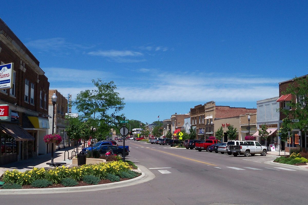 Main Street in downtown Brookings on a sunny day, with brick storefronts, parked pickups and a yellow flower bed on a curb island