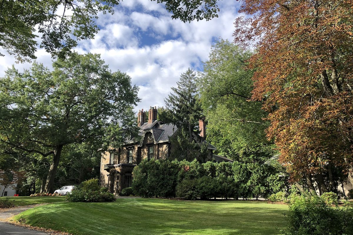A large Victorian stone house half hidden by tall trees and early fall foliage on Cottage Farm Road in Brookline, behind a wide green lawn