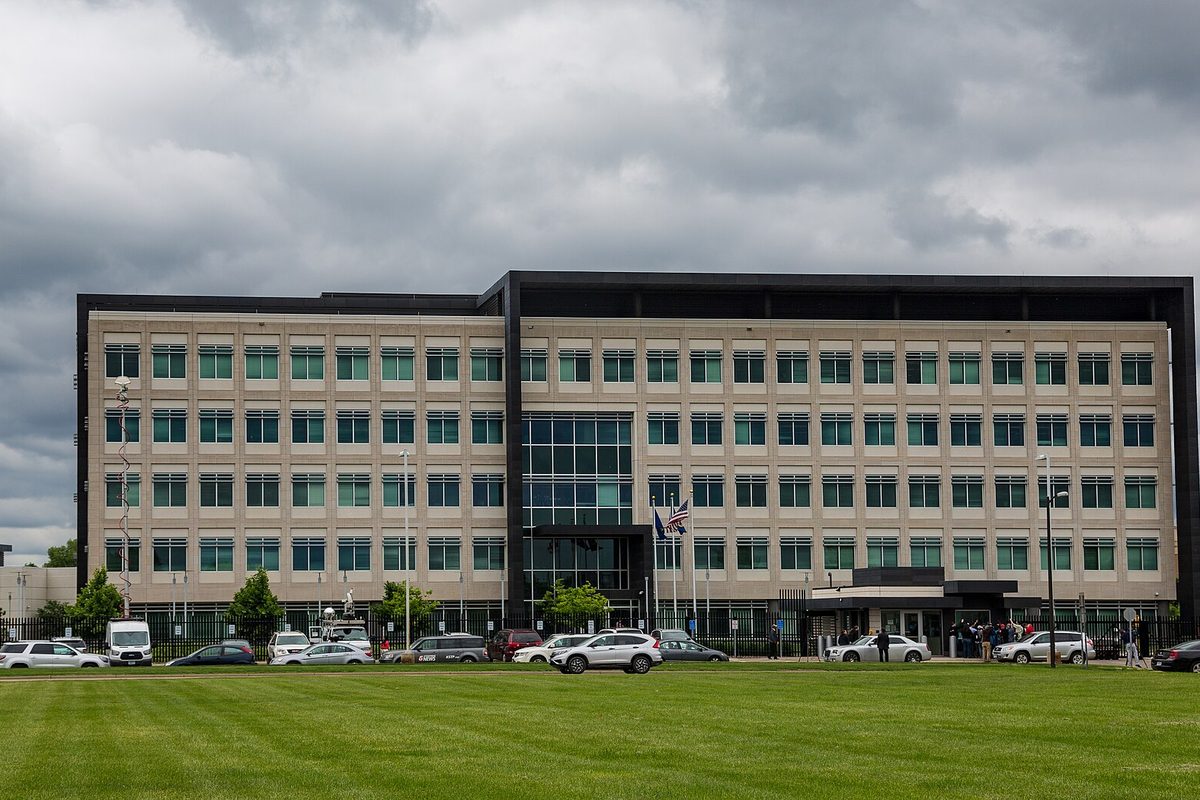 A six-story beige office building with rows of green-tinted windows on Freeway Boulevard in Brooklyn Center, cars parked along its front and a wide mowed lawn under a gray sky