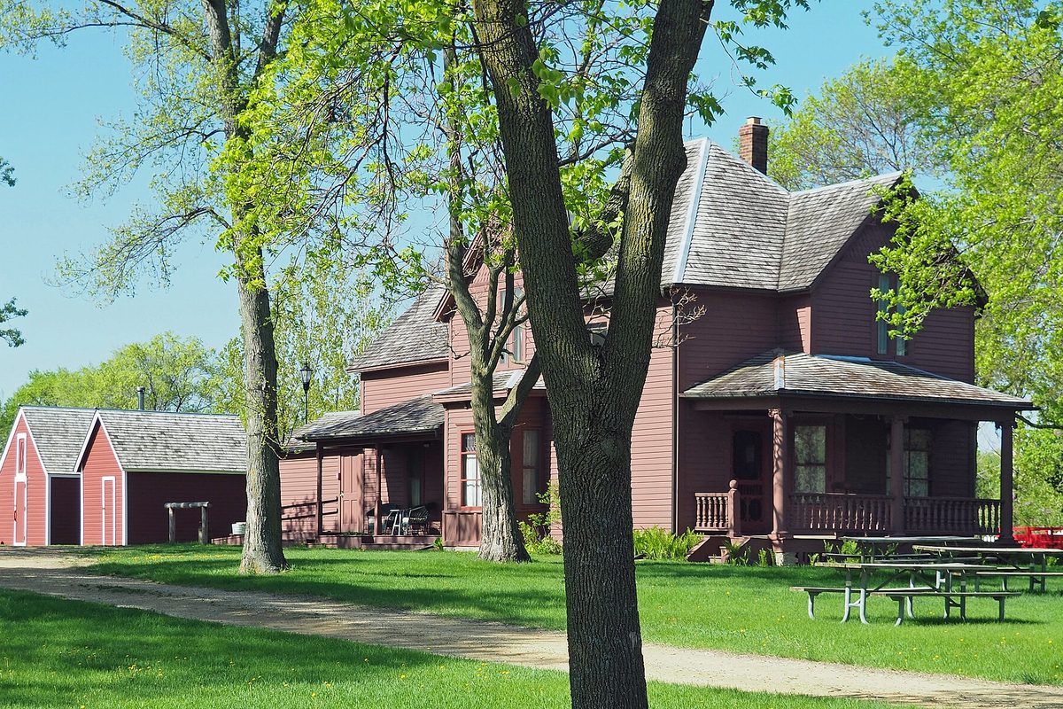 The red Eidem Homestead farmhouse and red outbuildings in Brooklyn Park, framed by spring trees, a gravel path and a green lawn