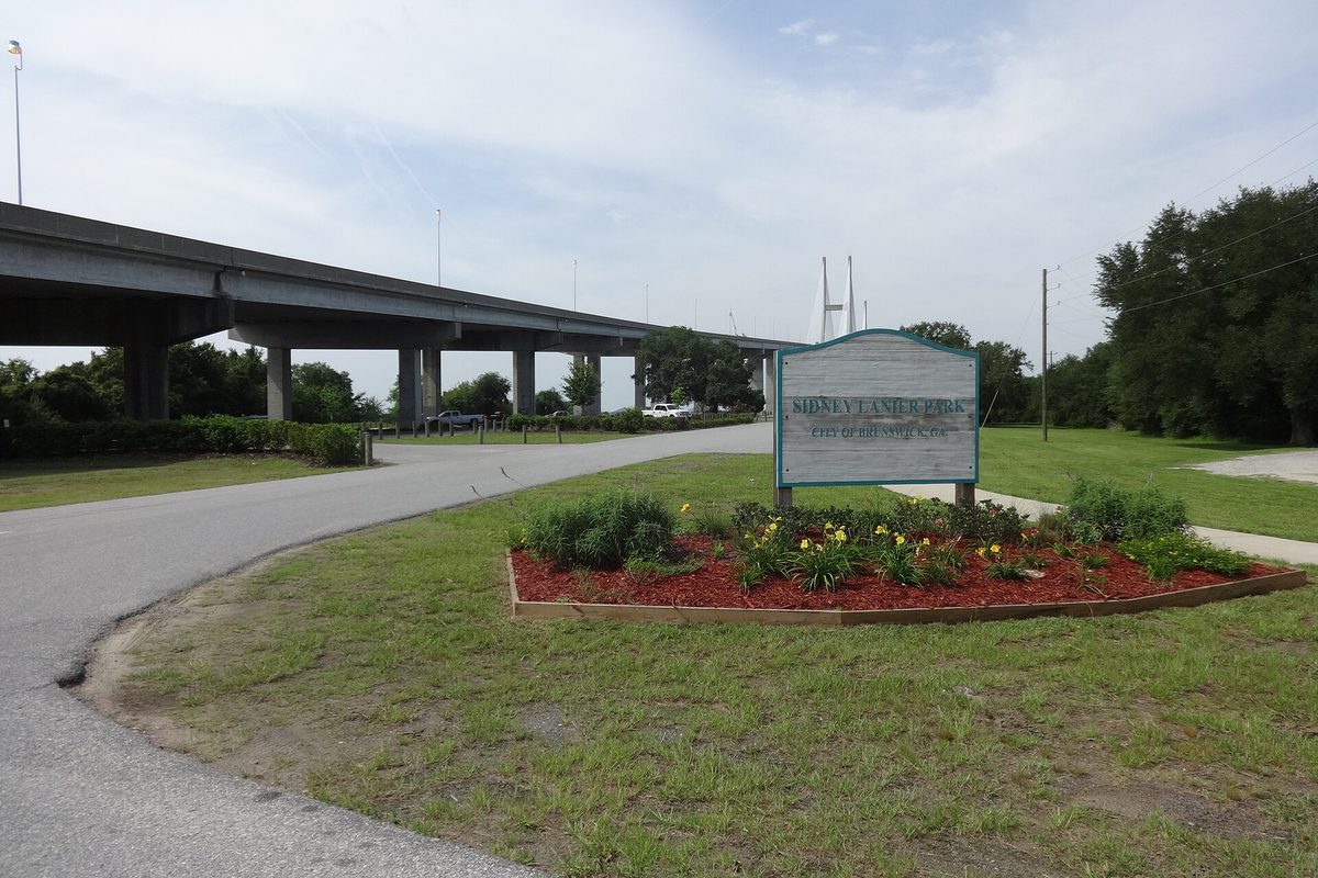 The Sidney Lanier Park sign and a flower bed beside a road, with an elevated highway approach and the towers of the Sidney Lanier Bridge behind