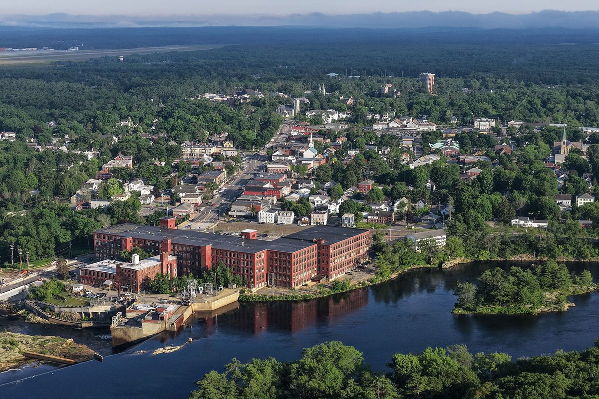 Aerial view of Brunswick with the long red brick former Cabot Mill on the river in the foreground, a dam at left, and the town's streets and steeples among trees behind