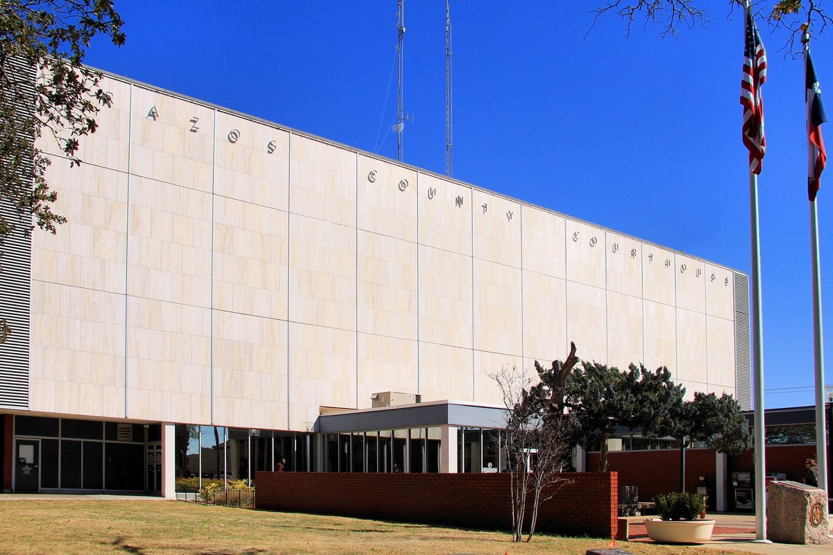 The pale stone Brazos County Courthouse in Bryan, its name spelled in spaced letters along the top, with U.S. and Texas flags flying under a deep blue sky