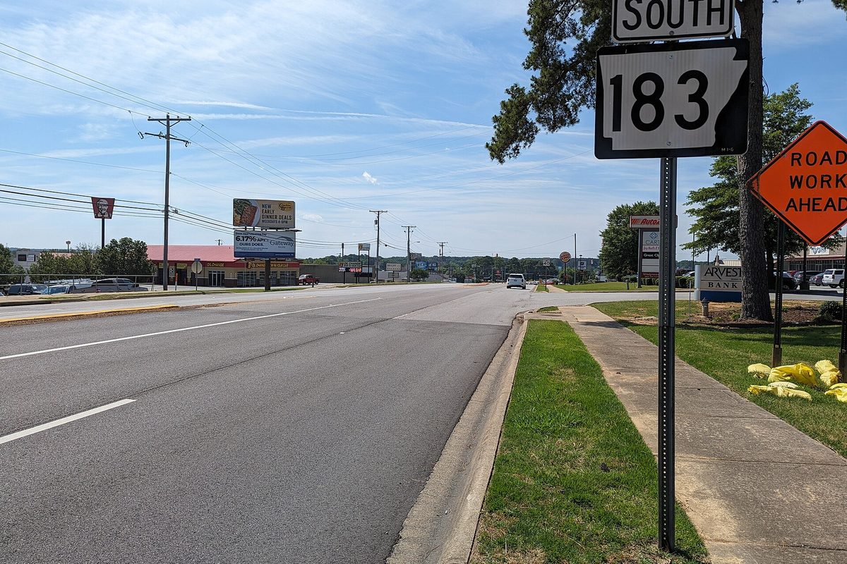 A South AR 183 route sign and a Road Work Ahead sign beside a wide four-lane road lined with billboards and businesses in Bryant