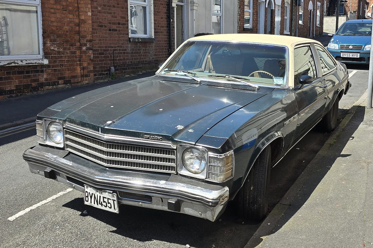 A dark green 1979 Buick Century fastback with a cream vinyl roof and chrome grille, parked on a narrow street of brick houses