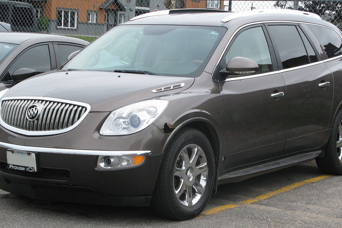 A dark brown first-generation Buick Enclave crossover with chrome grille and alloy wheels, parked by a chain-link fence