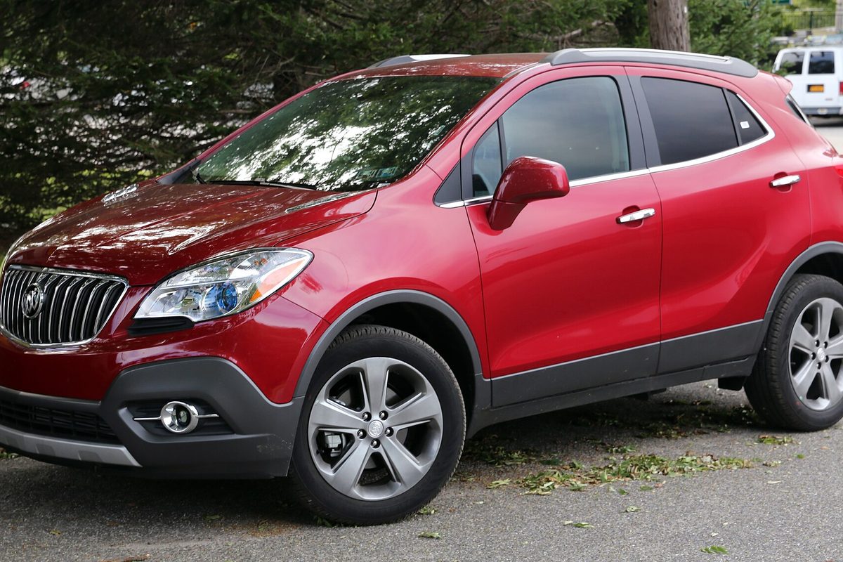 A dark red 2013 Buick Encore small SUV parked on pavement under evergreen trees, seen from the front left