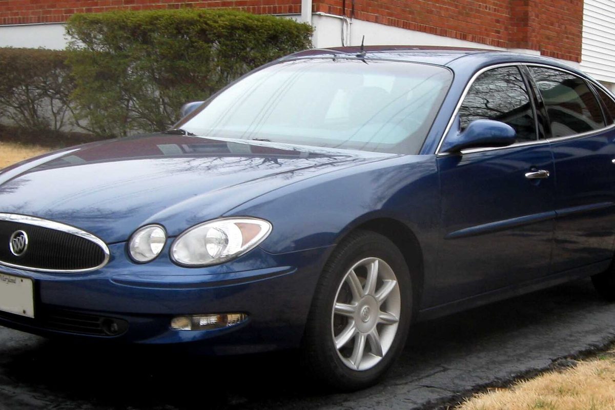 A dark blue 2006-2007 Buick LaCrosse sedan parked on a driveway beside a brick house and hedges