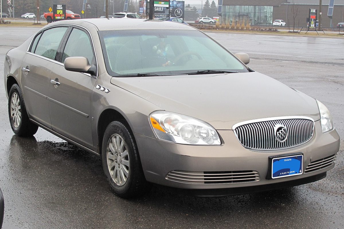 A tan metallic 2006 Buick Lucerne sedan with a chrome waterfall grille, parked on a wet lot on a gray day