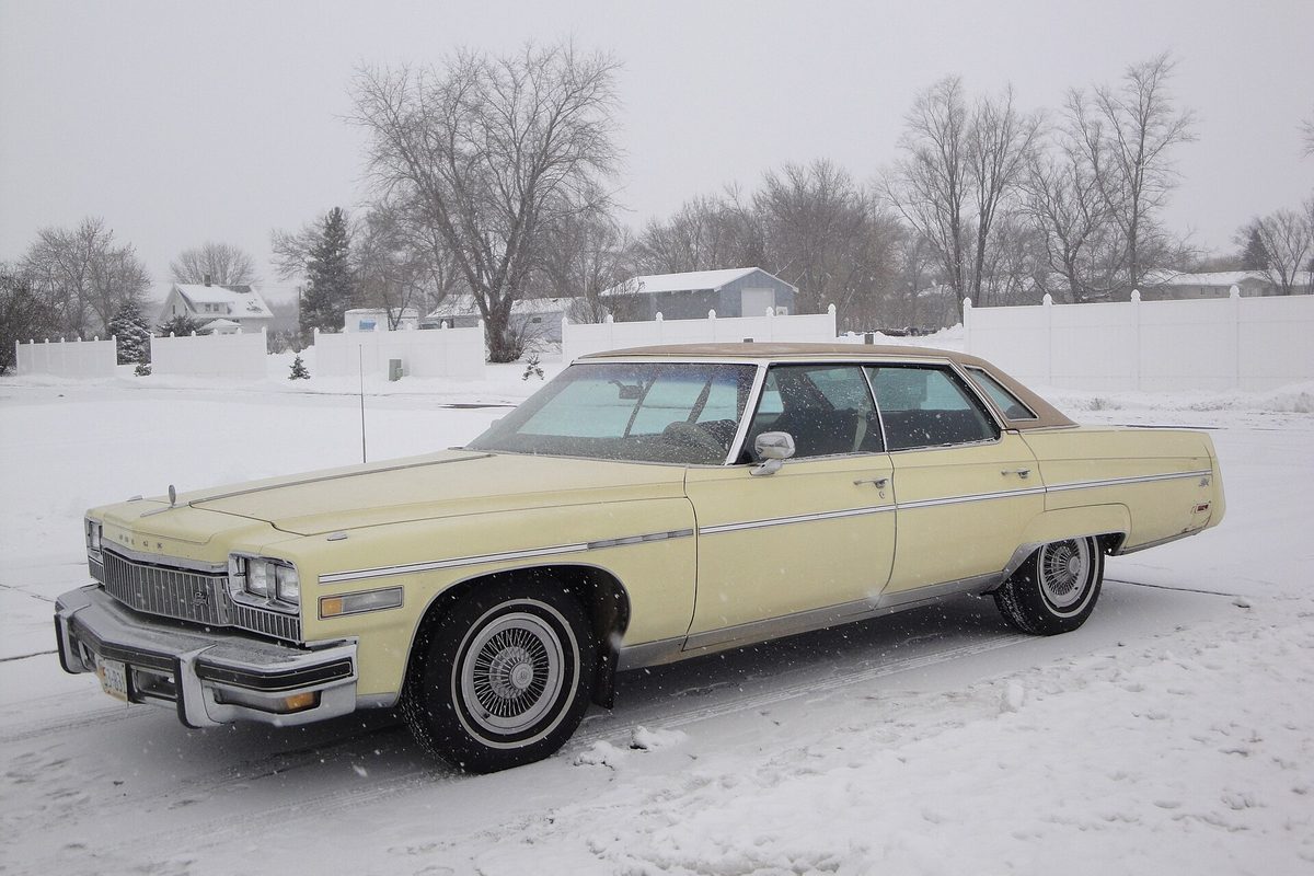 A pale yellow 1976 Buick Electra Park Avenue four-door hardtop with a brown vinyl roof, parked in falling snow