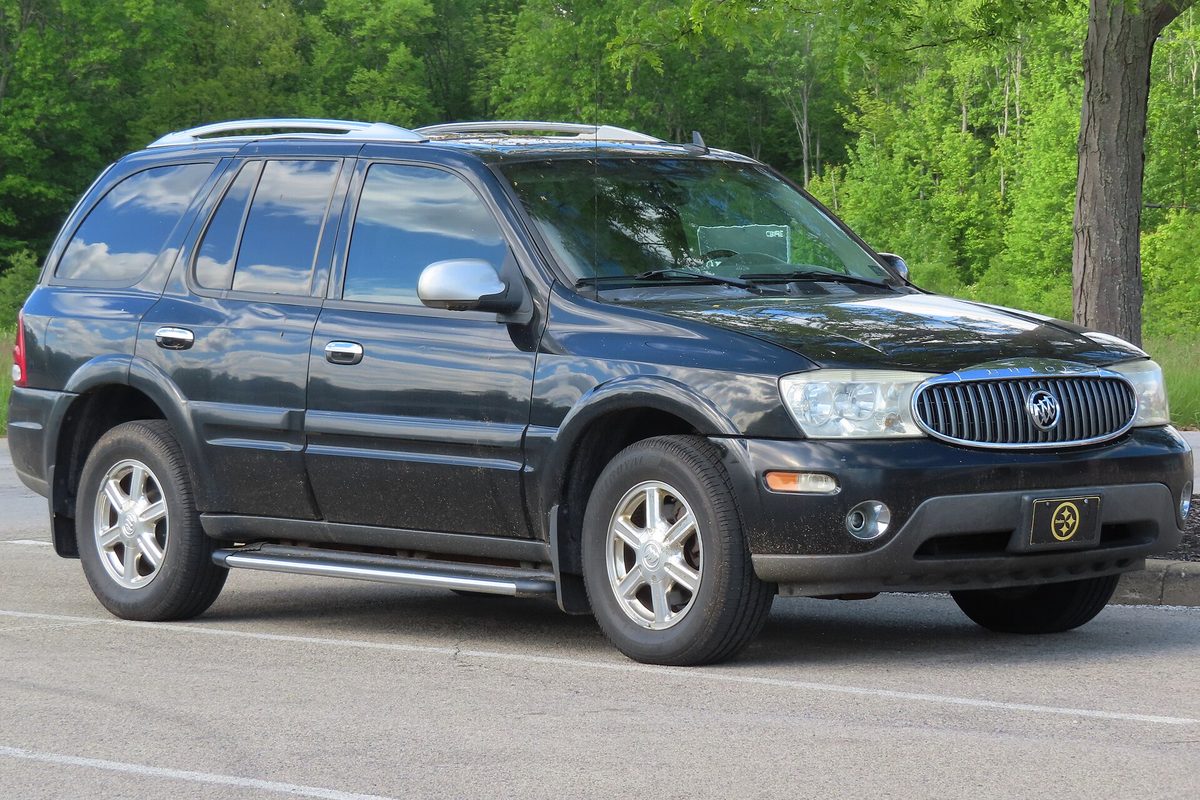 A black 2007 Buick Rainier SUV with a chrome grille and side steps, parked in a lot in front of green trees
