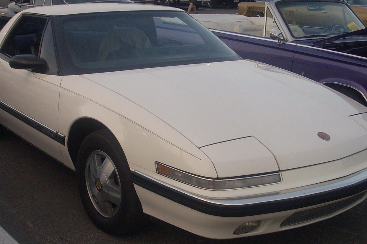A white Buick Reatta two-seat coupe seen from the front, parked in a lot beside a purple classic convertible