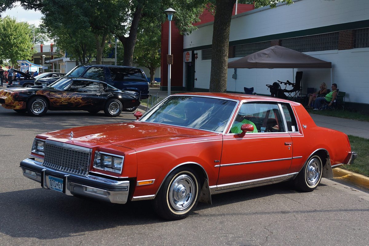 A red 1980 Buick Regal coupe with chrome bumpers and whitewall tires, parked at an outdoor car show