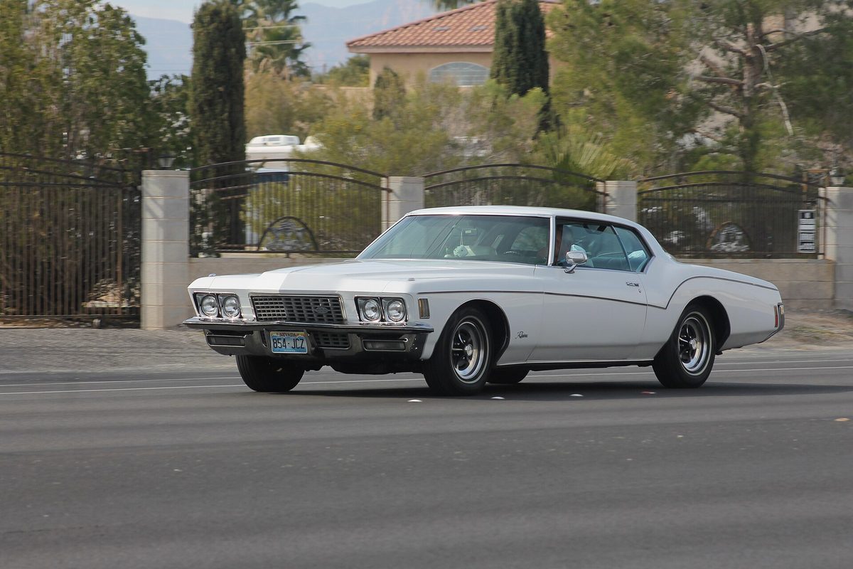 A white third-generation Buick Riviera two-door coupe driving on a road past a gated wall and palm trees