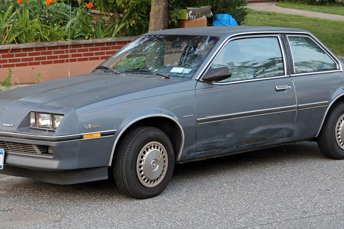 A gray Buick Skyhawk two-door coupe with a dented door, parked at a curb in front of a brick garden wall