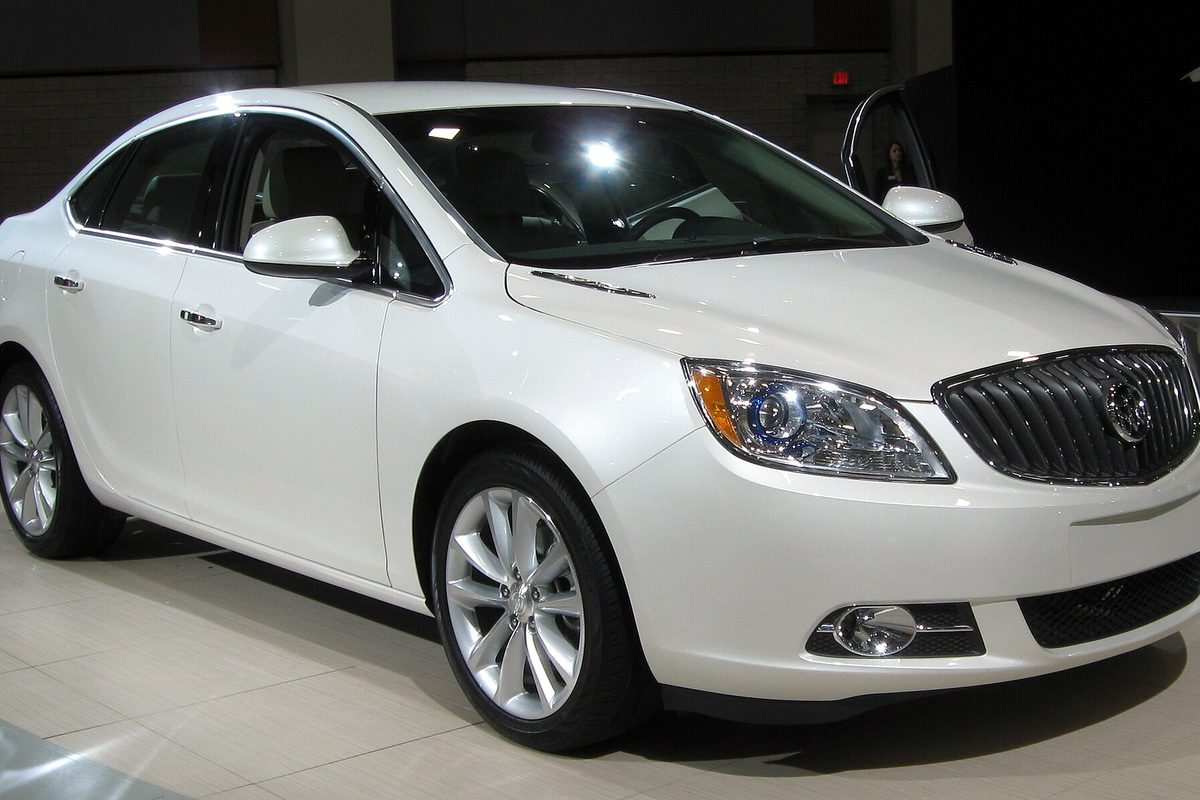 A pearl white 2012 Buick Verano sedan on a polished showroom floor, seen from the front right