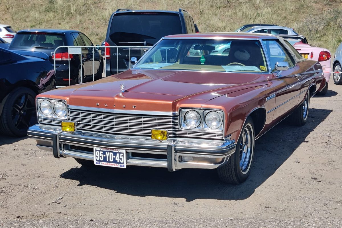 A copper-brown 1975 Buick LeSabre Custom Landau two-door with chrome bumpers and quad headlights, parked on a dirt lot beside newer cars