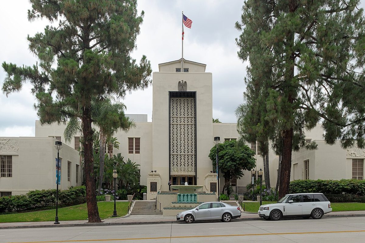 The cream-colored front of Burbank City Hall with its tall central tower and flag, framed by pines, with two cars parked at the curb