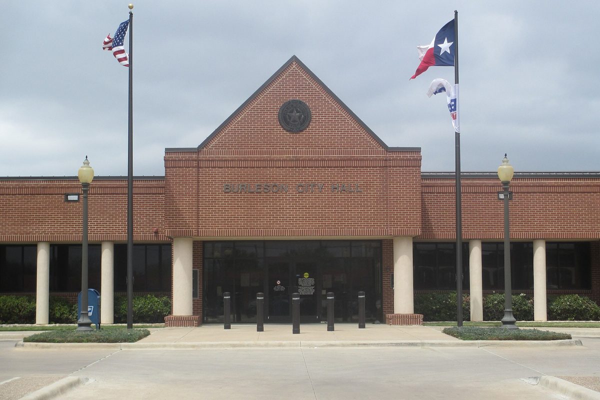 Burleson City Hall, a red-brick building with a peaked gable and a round seal over the entrance, flanked by U.S. and Texas flags on tall poles