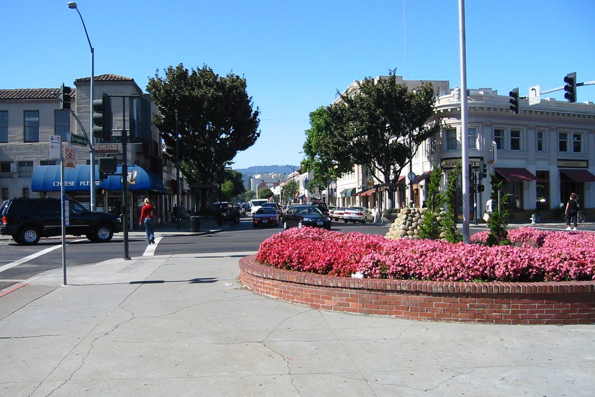 Looking west along Burlingame Avenue in the business district, with a brick planter of pink flowers, awninged storefronts and parked cars