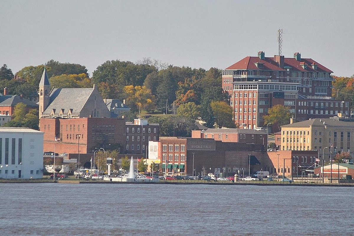 Downtown Burlington seen across the river, with brick buildings, a church steeple, a fountain at the water's edge and a large red-roofed brick building on the hill