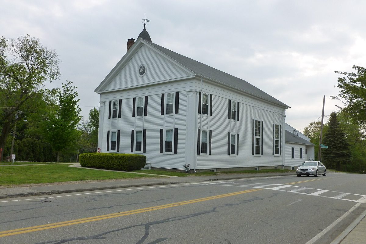 A white two-story clapboard church with black shutters and a small cupola on Lexington Street in Burlington, with a silver car passing on the road