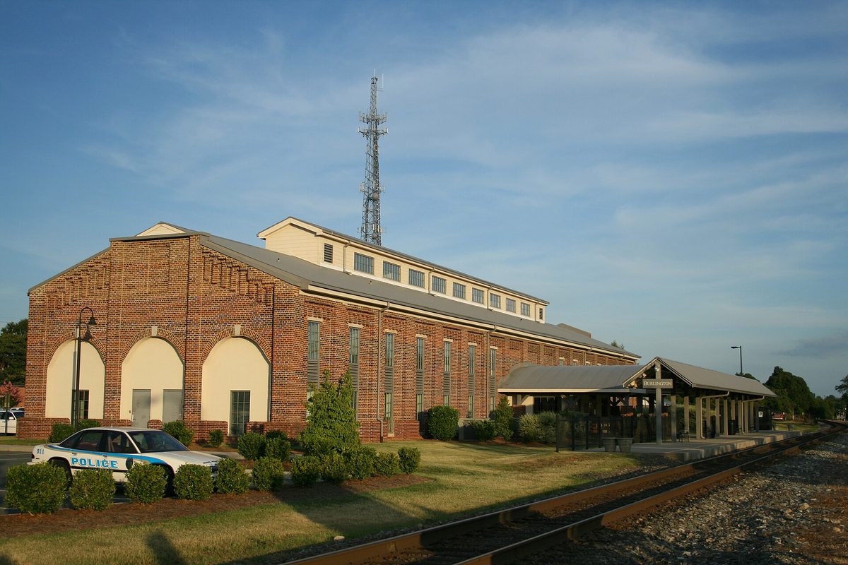 Burlington's brick train station with arched windows and a tall radio tower behind it, a police car parked out front and the tracks running past the platform
