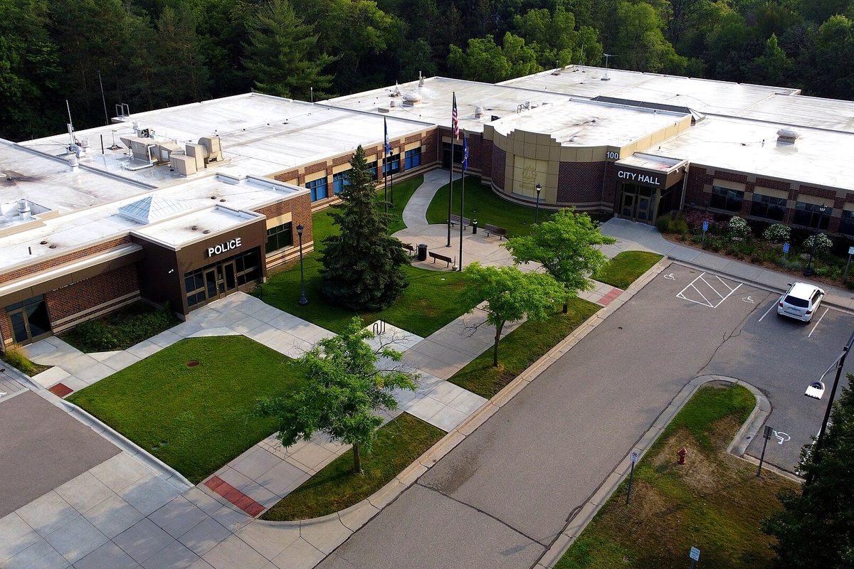 Aerial view of Burnsville City Hall and the police building, with flagpoles, lawns, young trees and a parking lot against a wooded hill