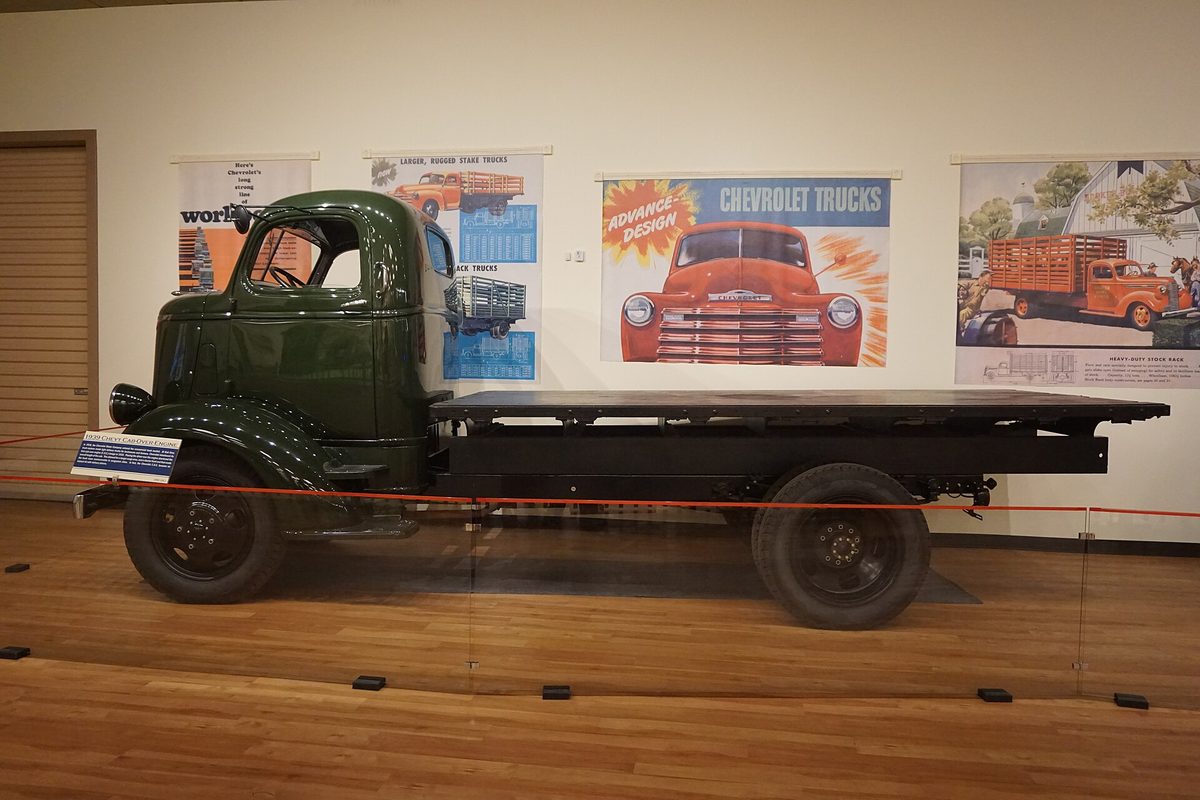 A dark green 1939 Chevrolet cab-over flatbed truck on display at the Sloan Museum at Courtland Center, with old Chevrolet truck posters on the wall behind it