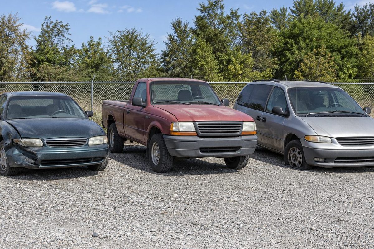 A dented blue sedan, a faded red pickup and a silver minivan parked side by side on a gravel lot by a chain-link fence